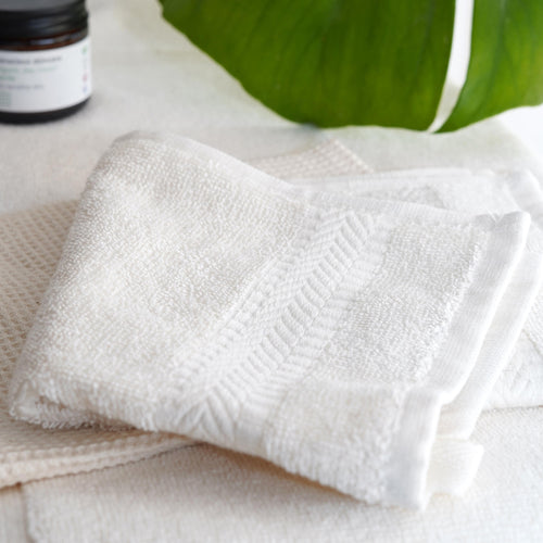 Organic cotton face cloth on white towel with jar and plant leaf in background